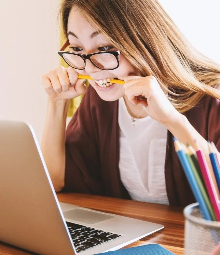 Junge Frau mit Brille sitzt vor einem Laptop, beißt in einen Bleistift und wirkt angespannt – Symbolbild für Stress, Deadline oder konzentriertes Arbeiten